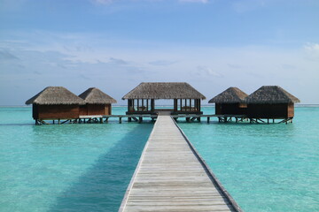 Tropical resort in Maldives showing overwater huts and bungalows with a long pier, thatched-roofs, crystal clear water and blue sky