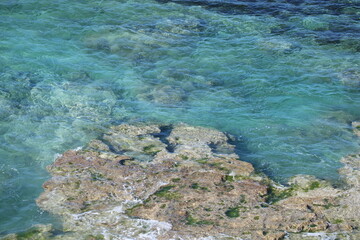Areal view of rocks and reef under crystal clear water, with some swell and surf going over the rocks