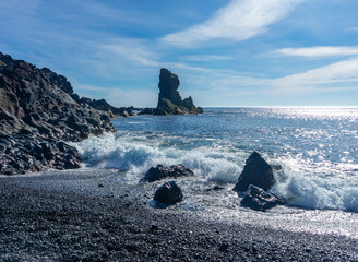 waves crashing on black sand beach with large rock in the distance