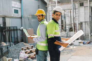 Contractor and workman wearing protective gear holds a laptop and a blueprint on a building site, Working Safety at Workplace Concept