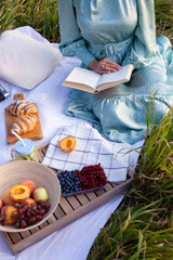 Fototapeta na wymiar Cropped photo of woman in long dress with short hair sitting on a white blanket with fruits and pastries and reading the book. Concept of having picnic in a city park during summer holidays or weekend