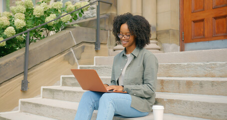 African American young woman, sitting outdoors with coffee, she works on the stairs, typing on her...