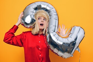 horizontal photo on a yellow background of a happy, joyful woman standing in a red shirt with a balloon in the form of the number two of silver color sticking her head into it
