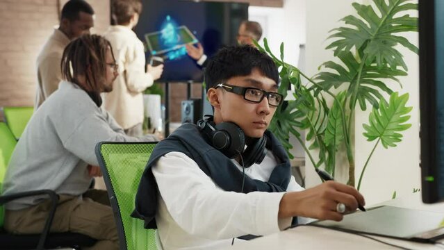 Asian male artist works in headphones in a studio for the development of computer games and applications. In the background, a team of developers are discussing a 3D model.