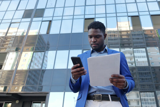 African American Realtor Checks Information In Paper Document With Phone. Man Entrepreneur Relates To Contract Of Apartment Purchase And Sale With Seriousness