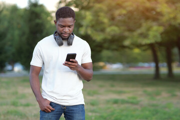 Fototapeta premium African American guy looks at smartphone and reads news update on Internet. Young man spends free time looking through social media in park at sunset, copyspace