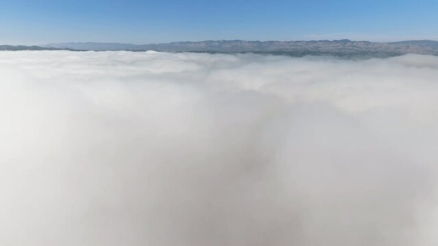 Drone shot of thick, milky clouds above the valley. Majestic blue skyline with mountain range in the background as seen from top. High quality 4k footage