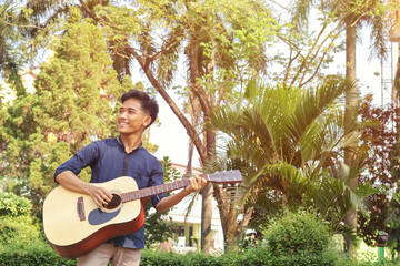 Portrait of attractive Asian man in casual shirt playing guitar while walking around in the park