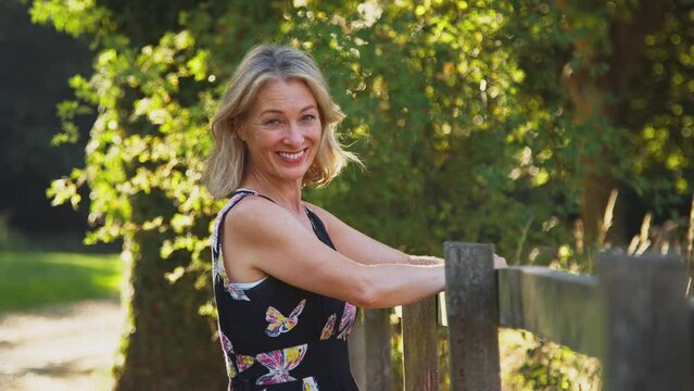 Casually dressed smiling mature woman leaning on wooden fence on walk in summer countryside - shot in slow motion