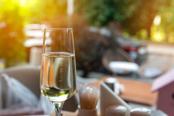 a glass of champagne is on the table of an outdoor cafe in the background is a park street, the greenery of trees and the setting sun. A relaxed and comfortable pastime - obrazy, fototapety, plakaty