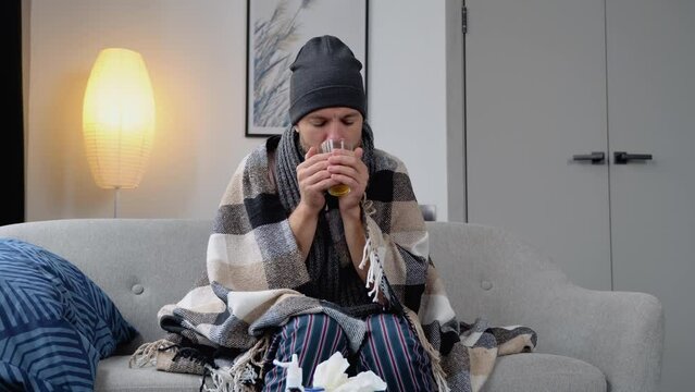 Health, flu and people concept - sick young man in hat wrapped in a blanket drinking hot tea at home. man warming his hands. Heating season