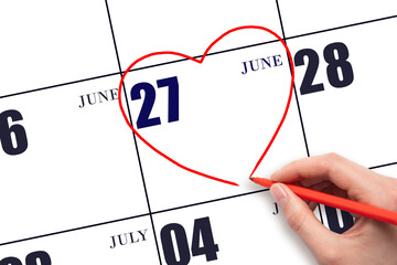 A woman's hand drawing a red heart shape on the calendar date of 27 June. Heart as a symbol of love.