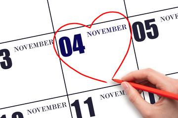 A woman's hand drawing a red heart shape on the calendar date of 4 November. Heart as a symbol of...