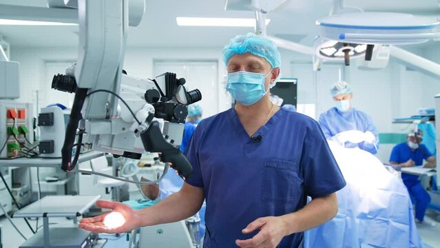 Demonstration of work and usage of advanced surgical equipment. Doctor shows the microscope placed in modern surgery room. Many doctors at backdrop in surgery.