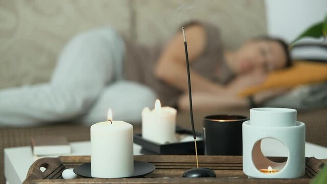 Close up of scented candles and aroma lamp. In the background woman is lying on sofa and sleeping, relaxing. Anti stress and holistic rest. Mental health concept.