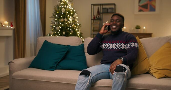 An African-looking student sits on sofa in the living room. Friend called him and wished Merry Christmas. The guy is very happy to communicate and smile. He is wearing warm sweater and ripped jeans.