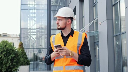 Designer engineer in protective helmet and vest standing on the background of a modern glass building uses the phone, examines the building project. Male professional reading email, message.