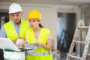 Man and woman construction workers in protection gear using laptop, holding documents ant talking on indoor building construction site