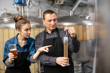 Fototapeta na wymiar Man brewmaster measuring beer sample with alcoholometer. His female colleague standing beside and pointing finger at graduated glass.