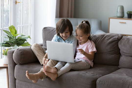 Two excited pretty gen Z kids using online learning app on laptop, talking on video call, watching content, browsing internet, playing video games, sitting on sofa at home alone