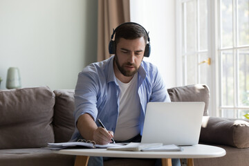 Focused serious millennial man in wireless headphones talking video call on laptop, writing notes, sitting on sofa at home, speaking, Adult student taking online educational learning course