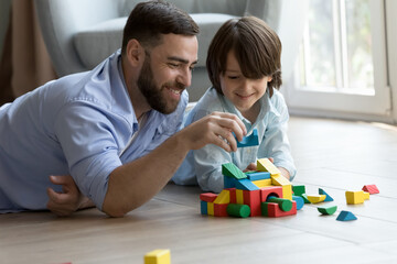 Happy dad teaching curious cute son kid to build toy forecast, castle, tower model from colorful blocks, playing learning games with child in heating floor. Fatherhood, family concept