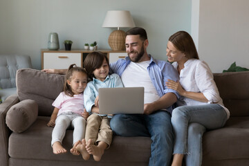 Happy family couple and cute cheerful kids using laptop on home couch, relaxing in living room...