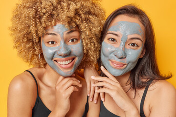 Gentle mixed race women touch jawline have glad expressions apply nourishing clay masks smile happily stand next to each other stand bare shoulders against yellow background. Wellness concept