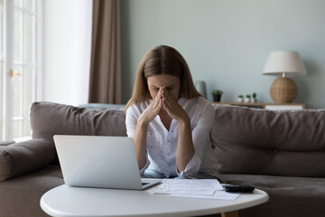 Desperate depressed millennial woman sitting at paper document, laptop, calculator at home, covering face with hands, feeling upset about lack of money, bankruptcy, job loss