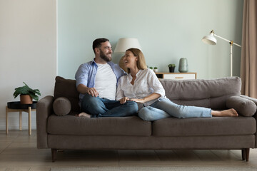 Happy joyful married couple resting on comfortable couch together, enjoying being at new cozy home, talking, laughing, having fun. Relationship, love, real estate for family concept - obrazy, fototapety, plakaty