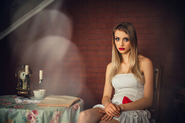 A gorgeous girl with bright make-up, with blond hair, in a light dress, sits in a room at a table. There is a candlestick with candles, a clock, a cup of coffee and a book on the table.