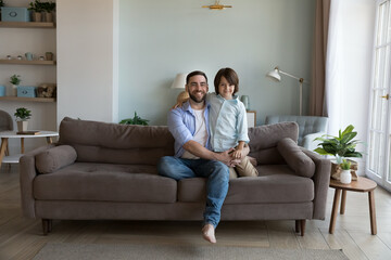 Happy handsome dad and little son kid sitting on sofa at home, hugging, looking at camera, enjoying...