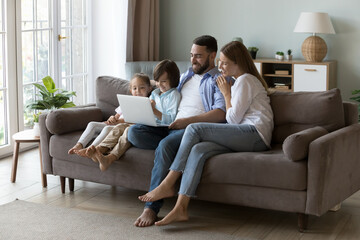 Happy millennial parents and two little gen Z resting on sofa, using laptop together, watching movie, interactive TV, talking on video call, chatting online. Family, Internet communication concept