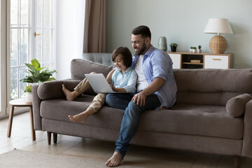 Happy millennial father hugging curious gen Z son kid, sitting on couch with laptop. Cheerful dad and little gamer boy playing virtual video game on computer. Fatherhood, family communication concept © fizkes