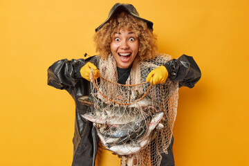 Amazed cheerful curly female fisher holds net full of big fish feels very happy to have successful fishing day dressed in black leather coat and hat isolated over yellow background. Big trophy