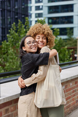 Vertical shot of happy diverse women glad to meet each other embrace and smile joyfully dressed in...