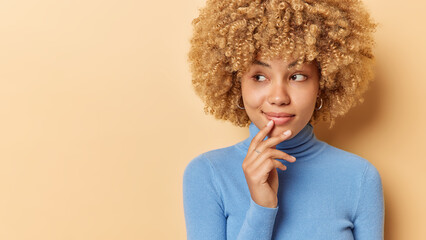 Studio shot of lovely curly haired young woman keeps hand near lips looks with curious expression aside daydreams about something dressed in casual blue turtleneck isolated over beige background