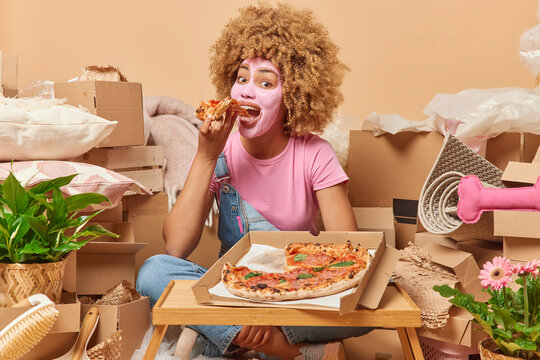 Positive woman with curly hair applies beauty mask on face eats delicious pizza celebrates moving day relocates to new apartment packs personal belongings in cardboard boxes. Relocation concept