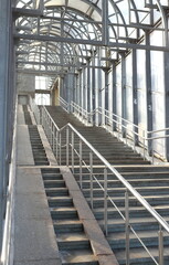 A staircase leading to a large covered bridge across the street. Perspective, arches, concrete, metal and glass.
