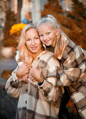 happy family, beautiful stylish blonde mother and daughter in warm beige checkered shirts walking in autumn city park. family look
