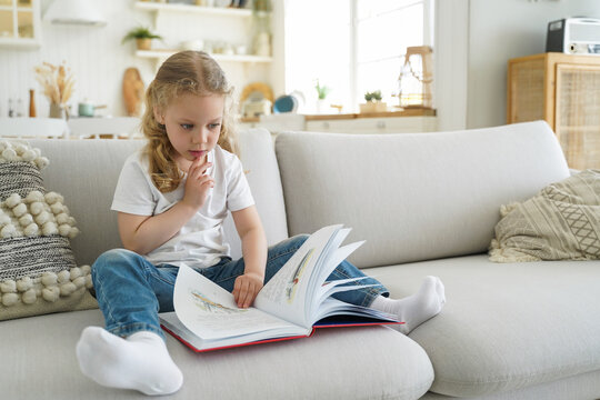 Pensive little child girl preschooler turns pages of fairytale book for reading, sit on sofa at home