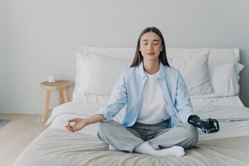 Disabled girl with bionic arm prosthesis practice yoga, meditating in lotus pose on bed at home