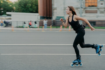 Horizontal shot of sporty energized woman enjoys rolleblading being photographed in motion poses on...