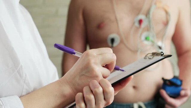 Close-up doctor's hand writing in patient's chart. Medical device for 24 hours daily Holter monitoring of electrocardiogram on a man's chest. Diagnosis of cardiovascular risk.