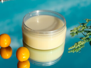 Cosmetic oil cream for moisturizing and nourishing the skin, isolated on a blue mirror background with a plant and a yellow plum. Skin care close-up.
