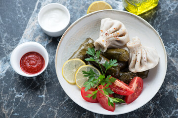 Plate with dolmades, khinkali dumplings, tomatoes, lemon and dips on a black marble background, horizontal shot