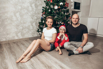 Family - mom, dad and son in Santa costume sitting under the Christmas tree on the floor in their apartment, looking into the camera