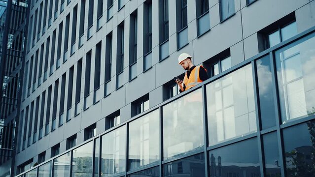 Professional architect engineer standing on the terrace of a modern building using mobile phone, reading message answering outdoors wearing in a safety helmet at a building construction site. 