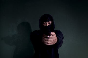 Criminal man in black hood holding a gun pointing victim, mask thief robber with his gun on hands...