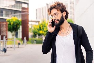 stylish young man with beard talking on mobile phone in the city, concept of urban lifestyle and technology of communication, copy space for text